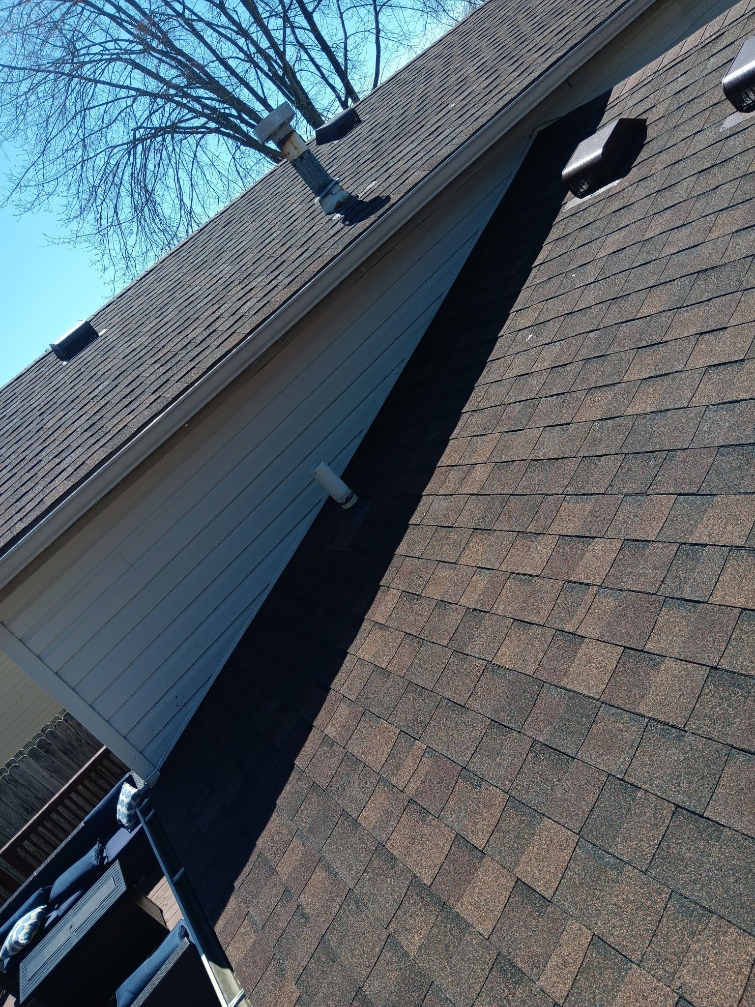A brown shingled roof with white siding, a vent pipe, and a tree against a blue sky.