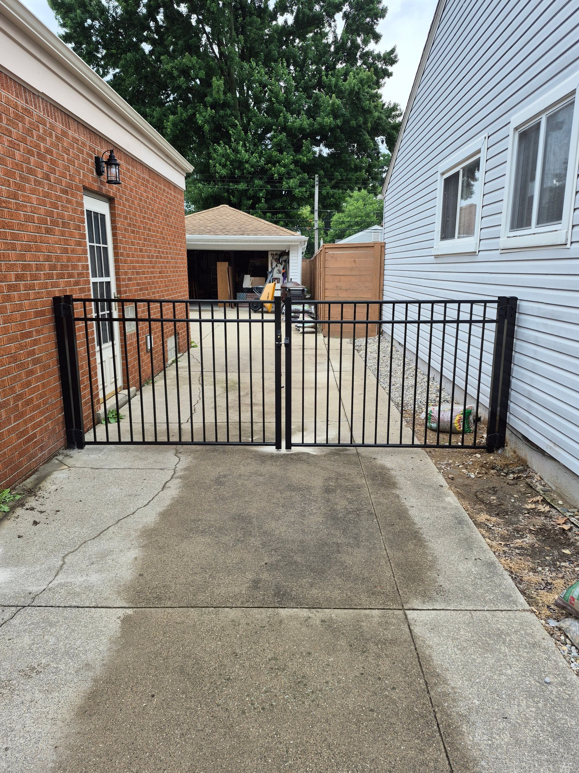 Black metal gates on a concrete driveway, between a brick building and a white house.
