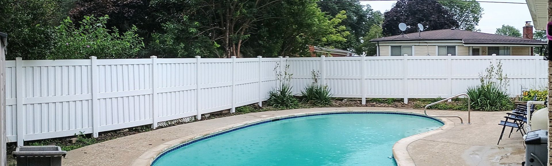 A backyard with a swimming pool, white fence, trees, and a house.