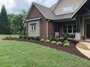 A brick house with stone accents, a covered front porch, and a landscaped garden bed with mulch and small green shrubs.