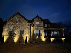 A large two-story stone house at twilight, illuminated by warm outdoor accent lighting against a dark blue sky.