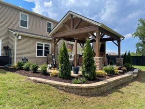 A wooden patio pavilion with stone pillars sits on a raised bed with shrubs and mulch beside a two-story beige house.