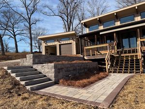 Stone steps lead up to a tiered retaining wall and a modern house with a wooden deck and garage under a blue sky.