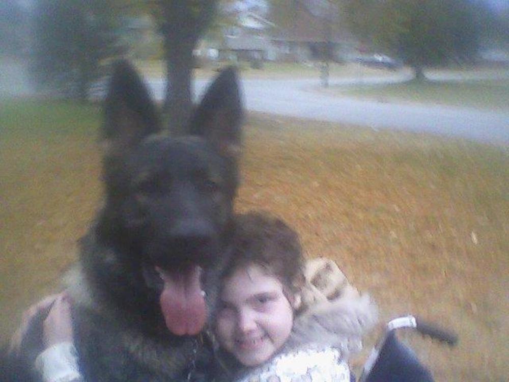 A girl in a wheelchair is hugging a large black dog.