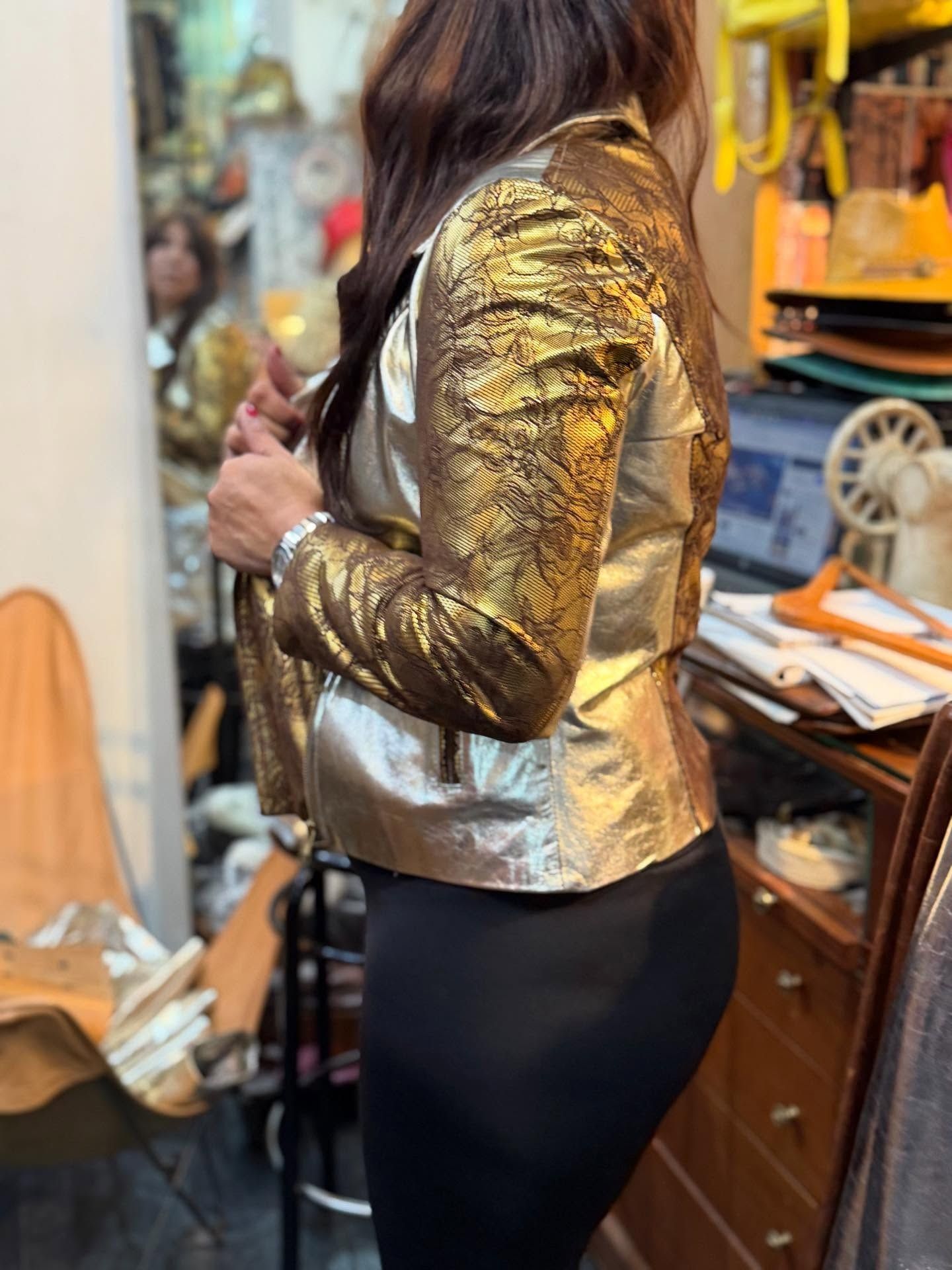 Mujer con chaqueta dorada y leggings negros posa de perfil derecho. Interior con materiales de costura.