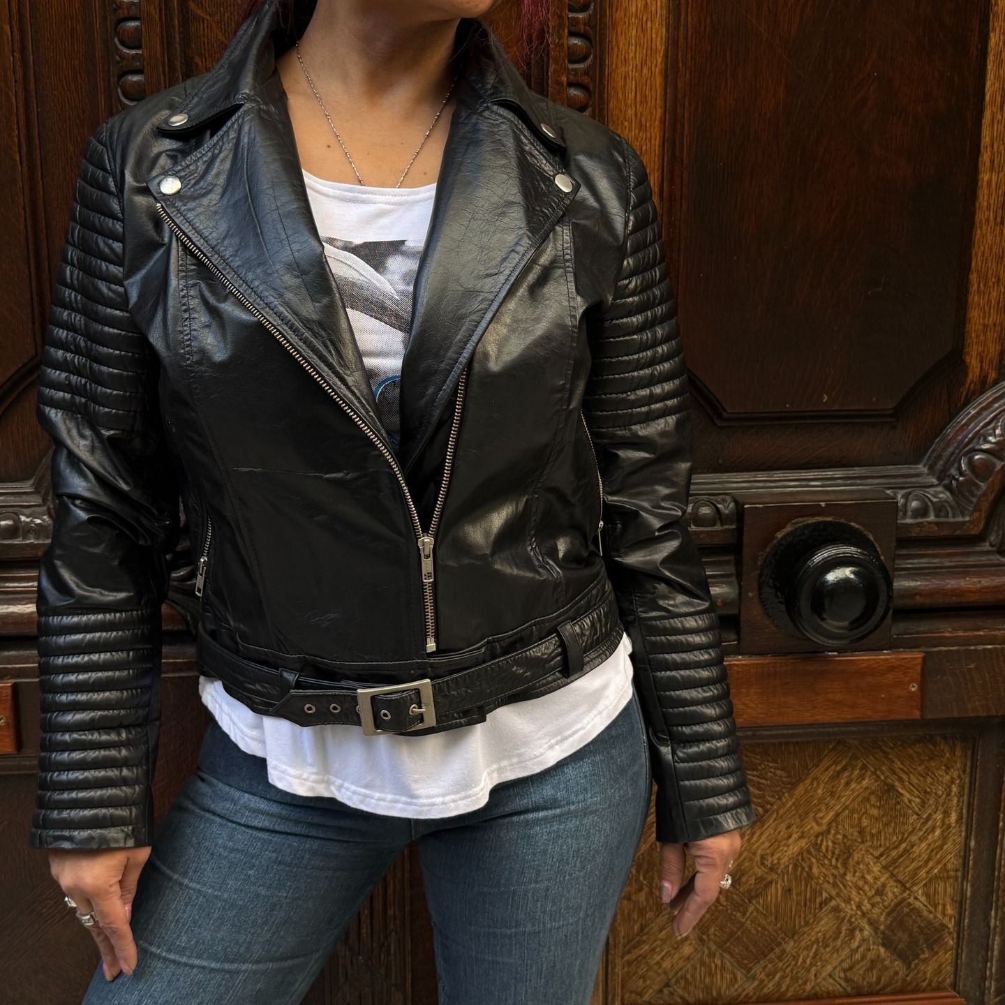 Mujer con chaqueta de cuero negra, camisa blanca y jeans, posando frente a una puerta de madera.