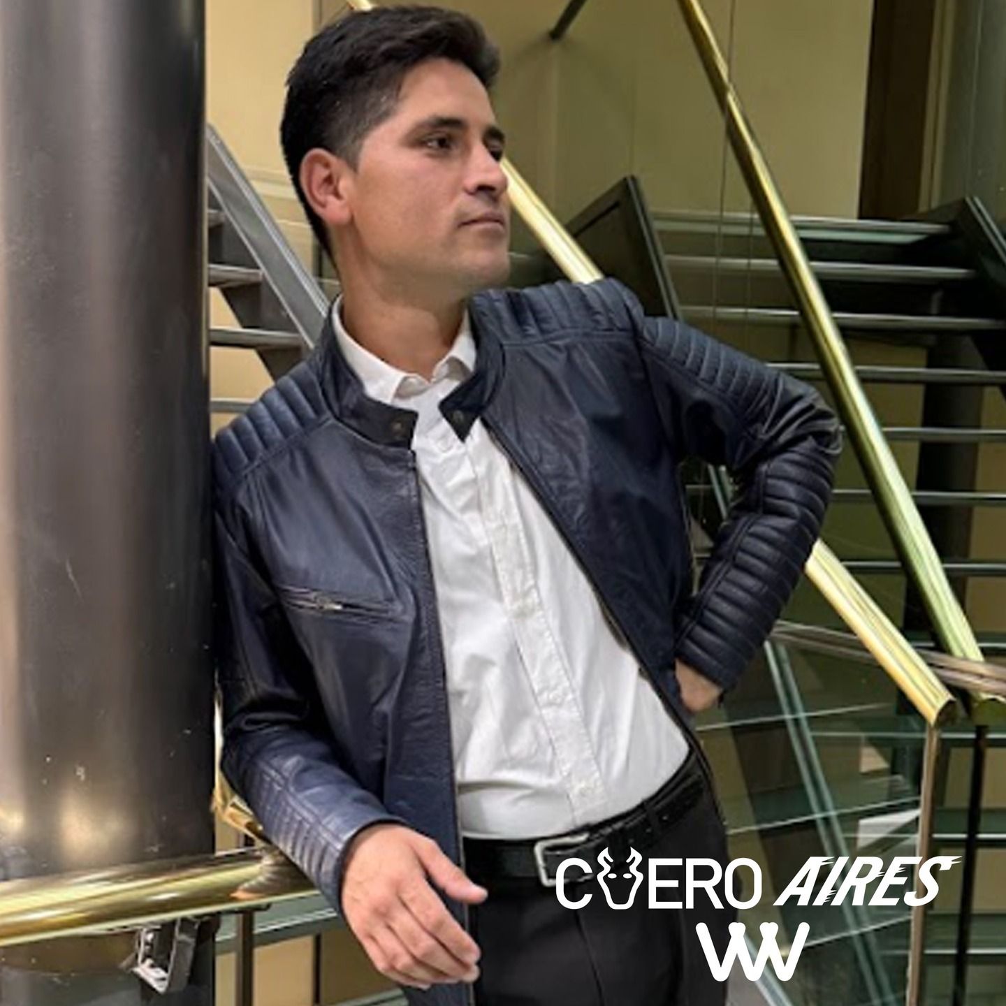 Un hombre con una chaqueta de cuero azul marino y una camisa blanca se apoya en una barandilla.