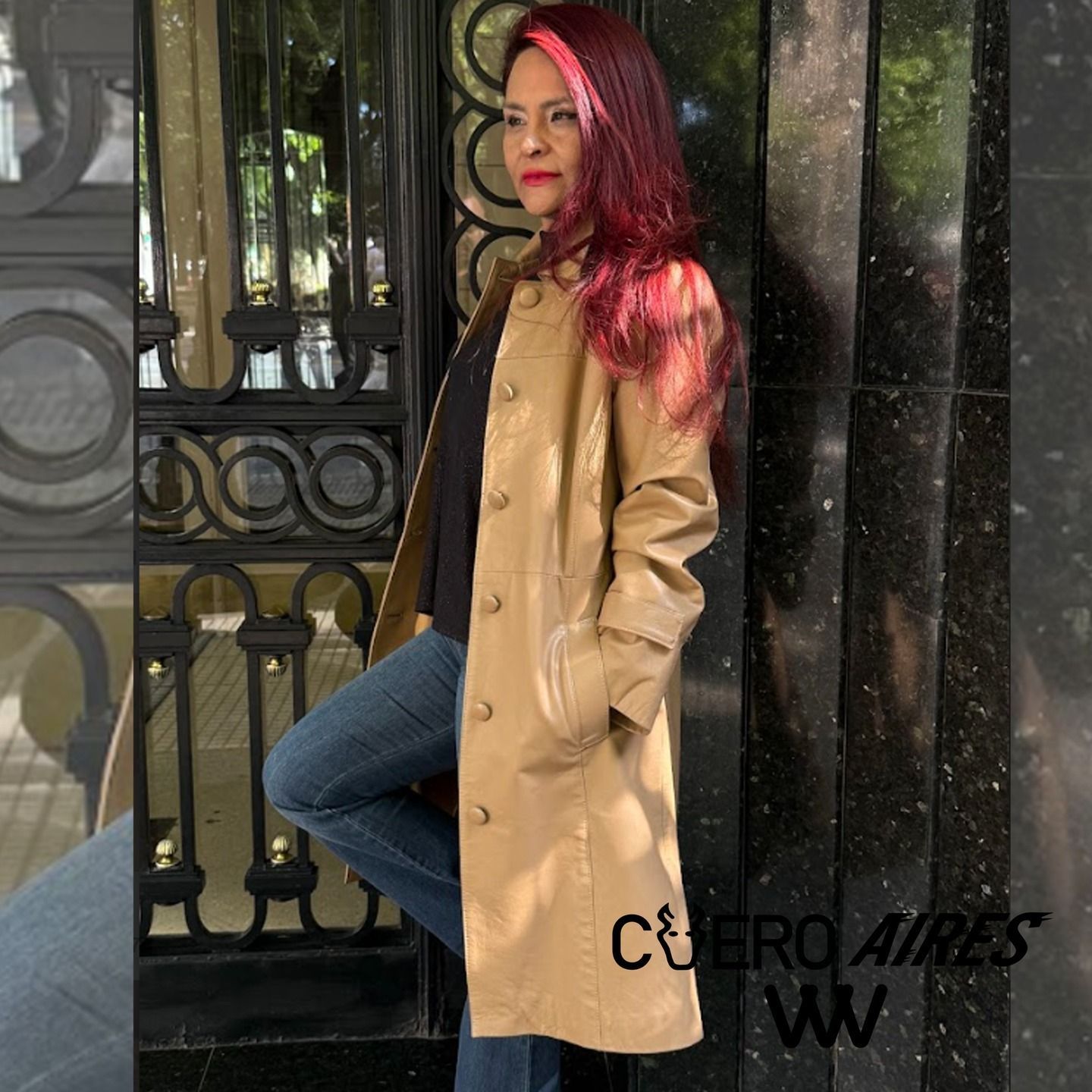 Mujer con abrigo color canela, apoyada contra una puerta oscura, vestida con jeans y una camisa negra.