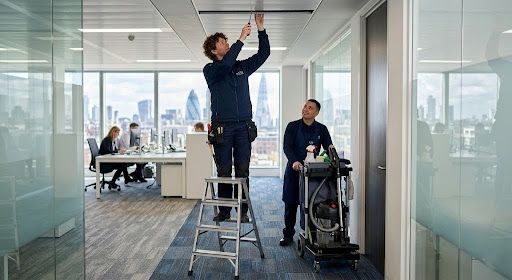 Two workers in a glass-walled office, one on a ladder adjusting ceiling lights, another beside equipment cart.