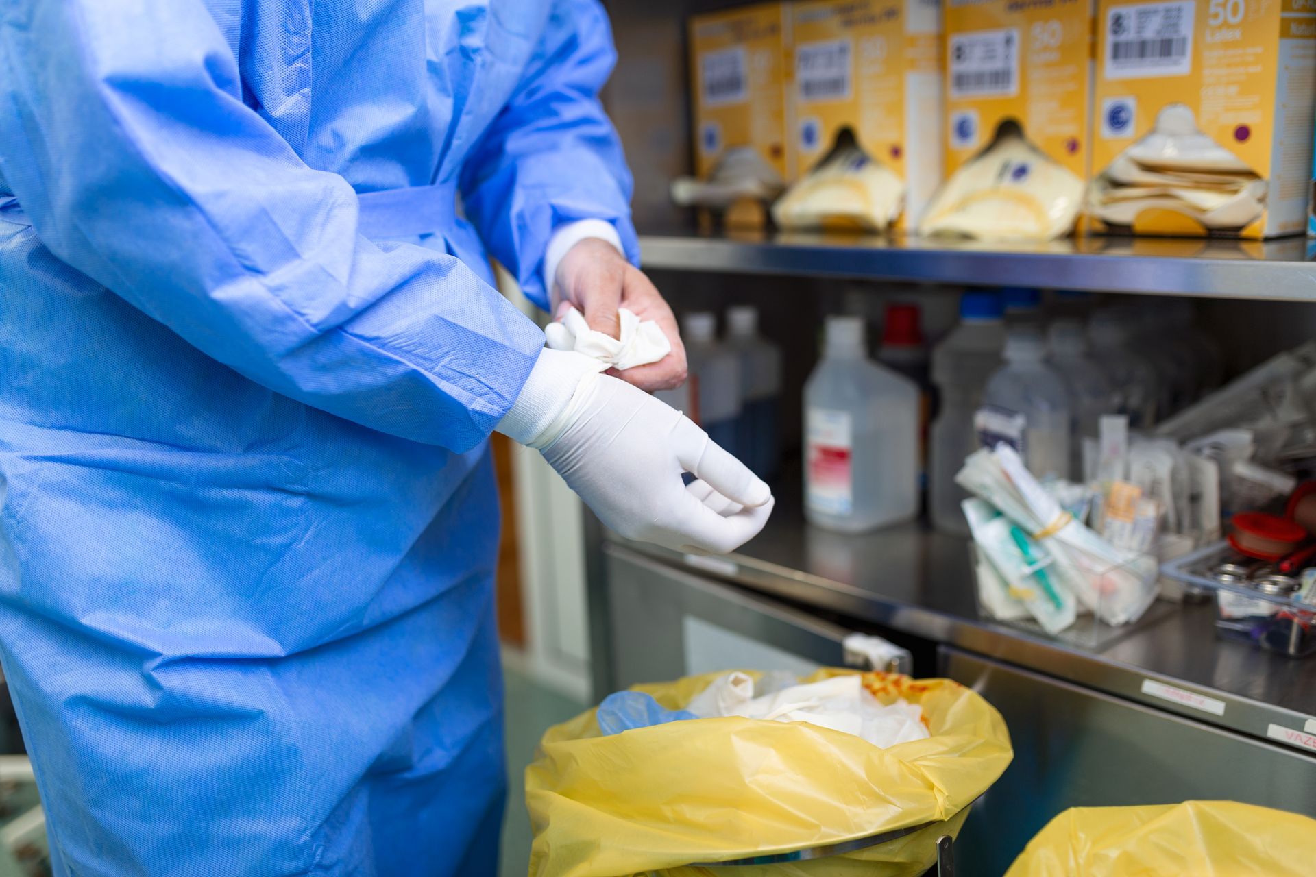 Person in blue medical suit putting on gloves in a medical setting, near a waste bin and supplies.