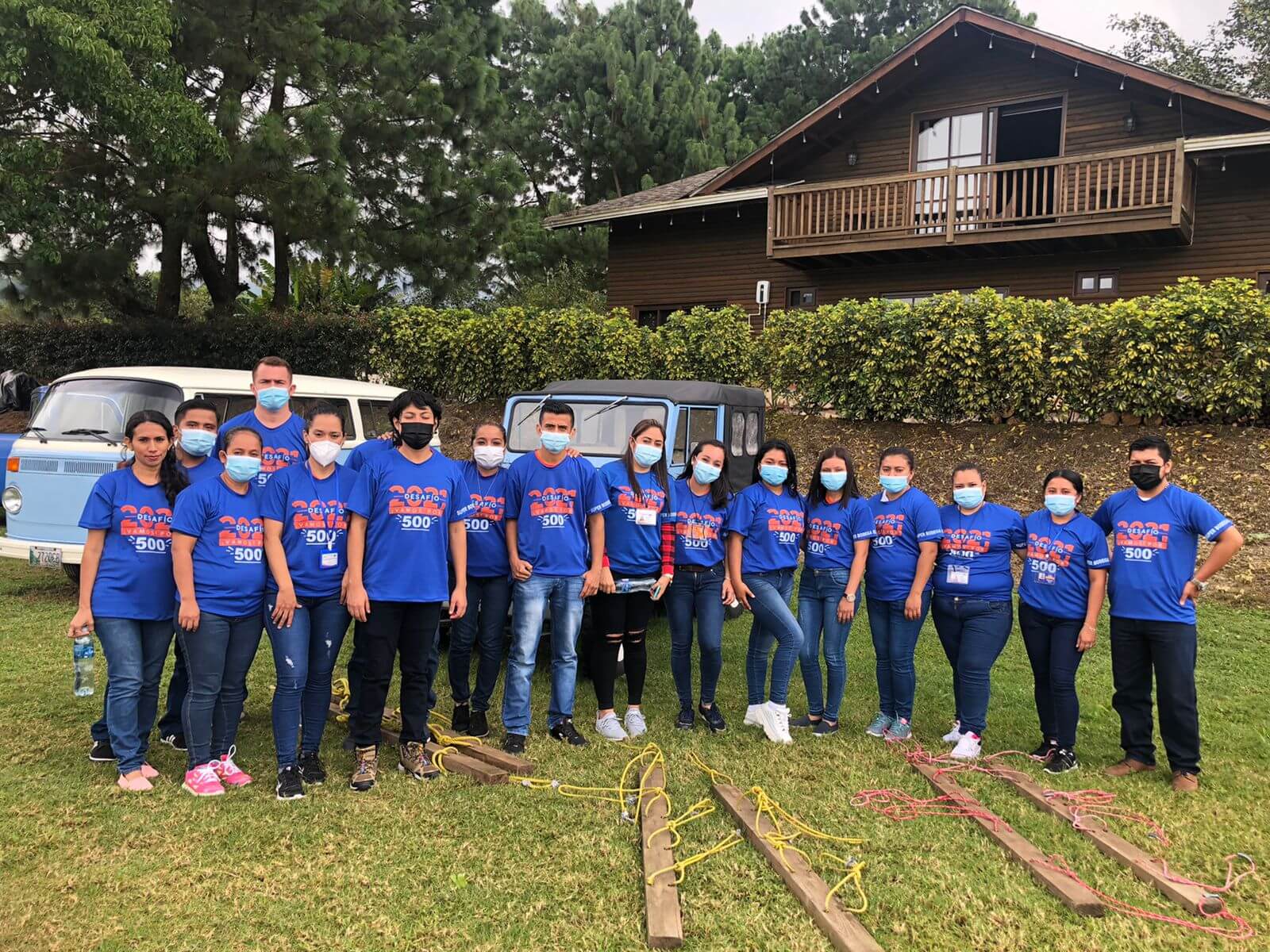 Grupo de personas con camisas azules posan al aire libre con una cabaña y vehículos.