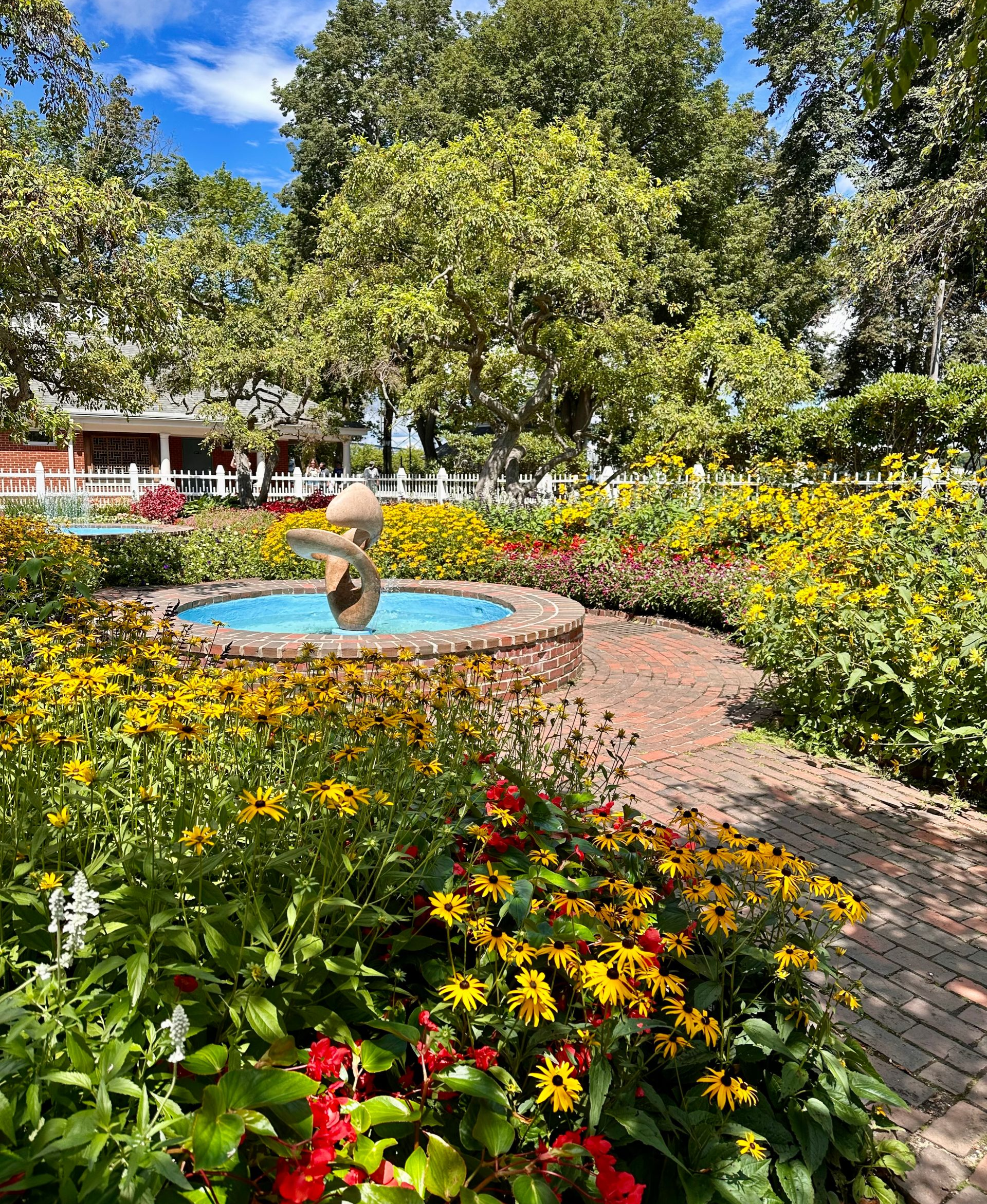 Sunny garden with a small fountain, brick path, and colorful flowers under leafy trees