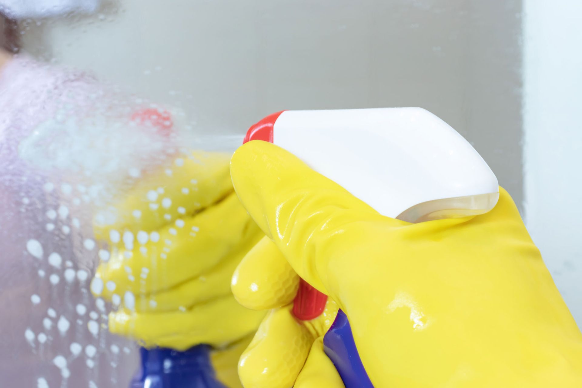 A person wearing yellow gloves is cleaning a mirror with a spray bottle.