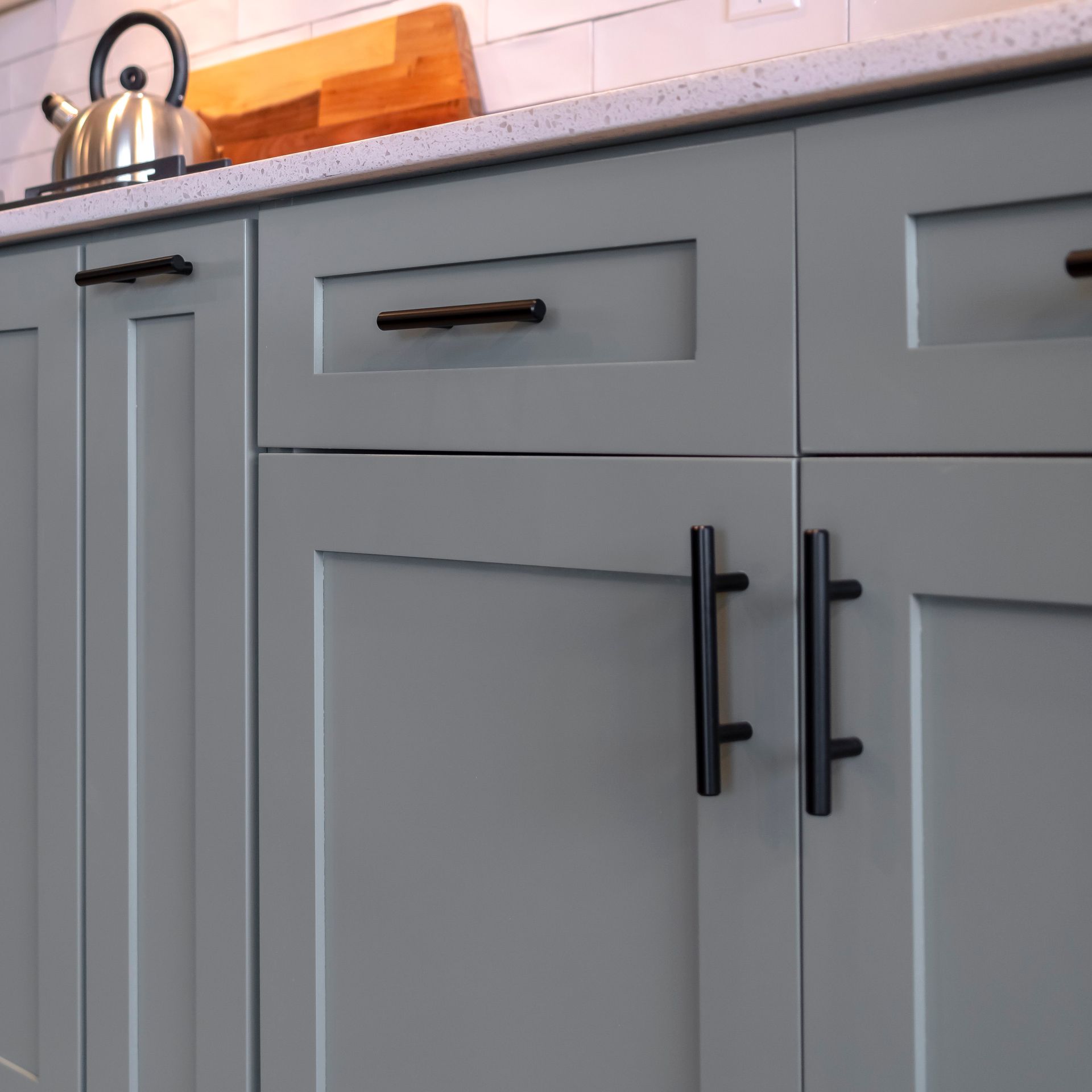 Gray kitchen cabinets with black handles; a kettle and cutting board are in the background.