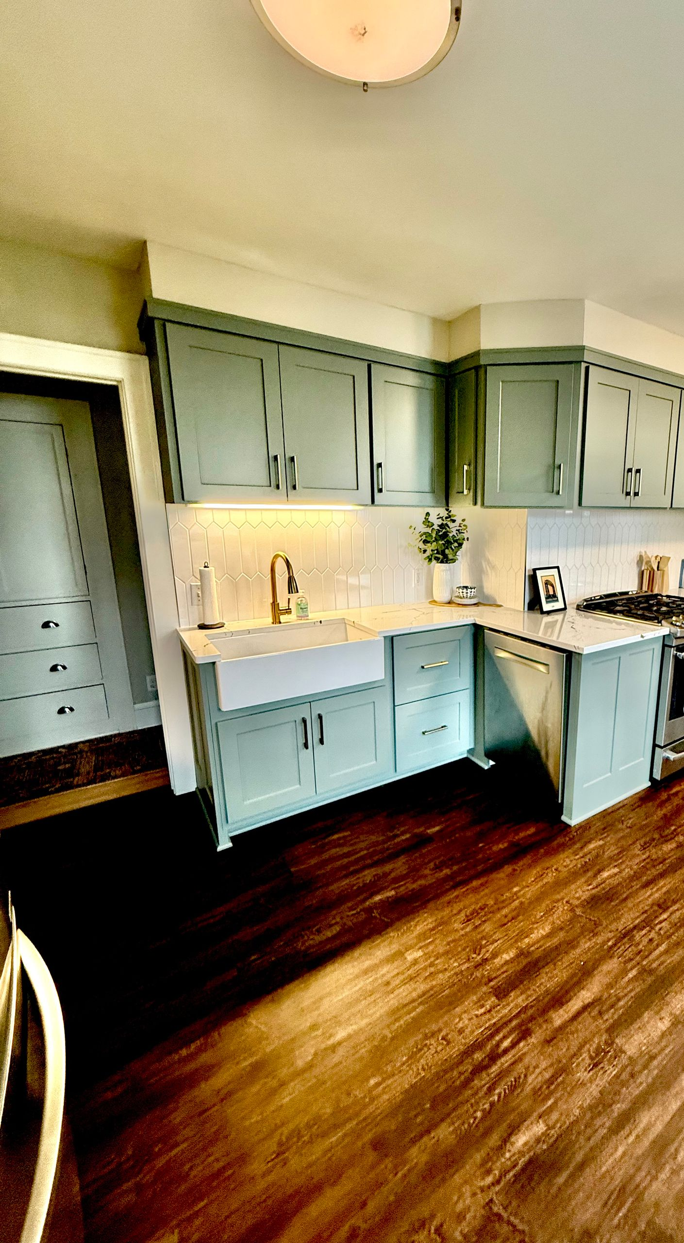 Kitchen with sage green cabinets, white backsplash, farmhouse sink, and wood flooring.