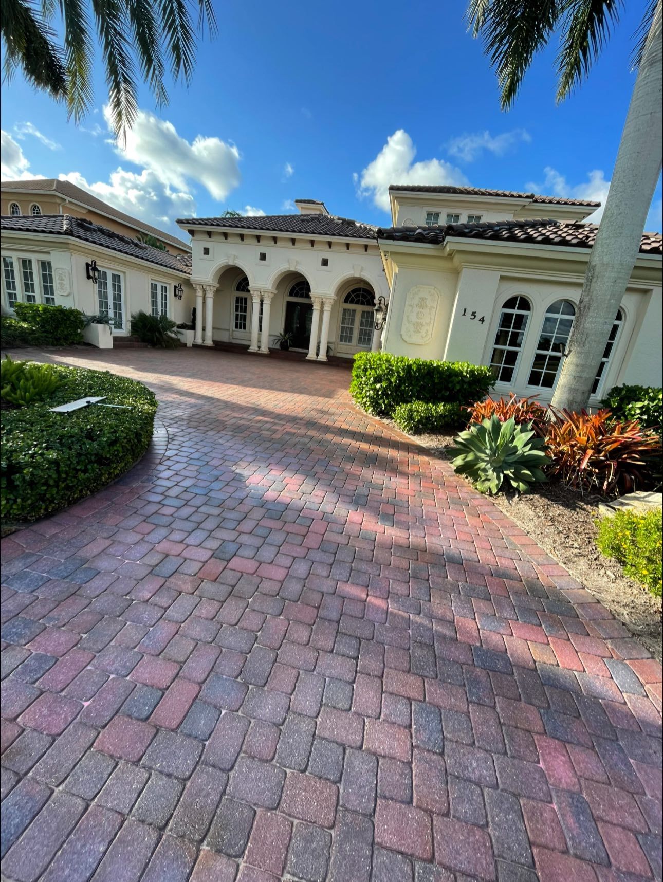 Brick driveway leading to a cream-colored Mediterranean-style house with arched doorways and palm trees against a blue sky.