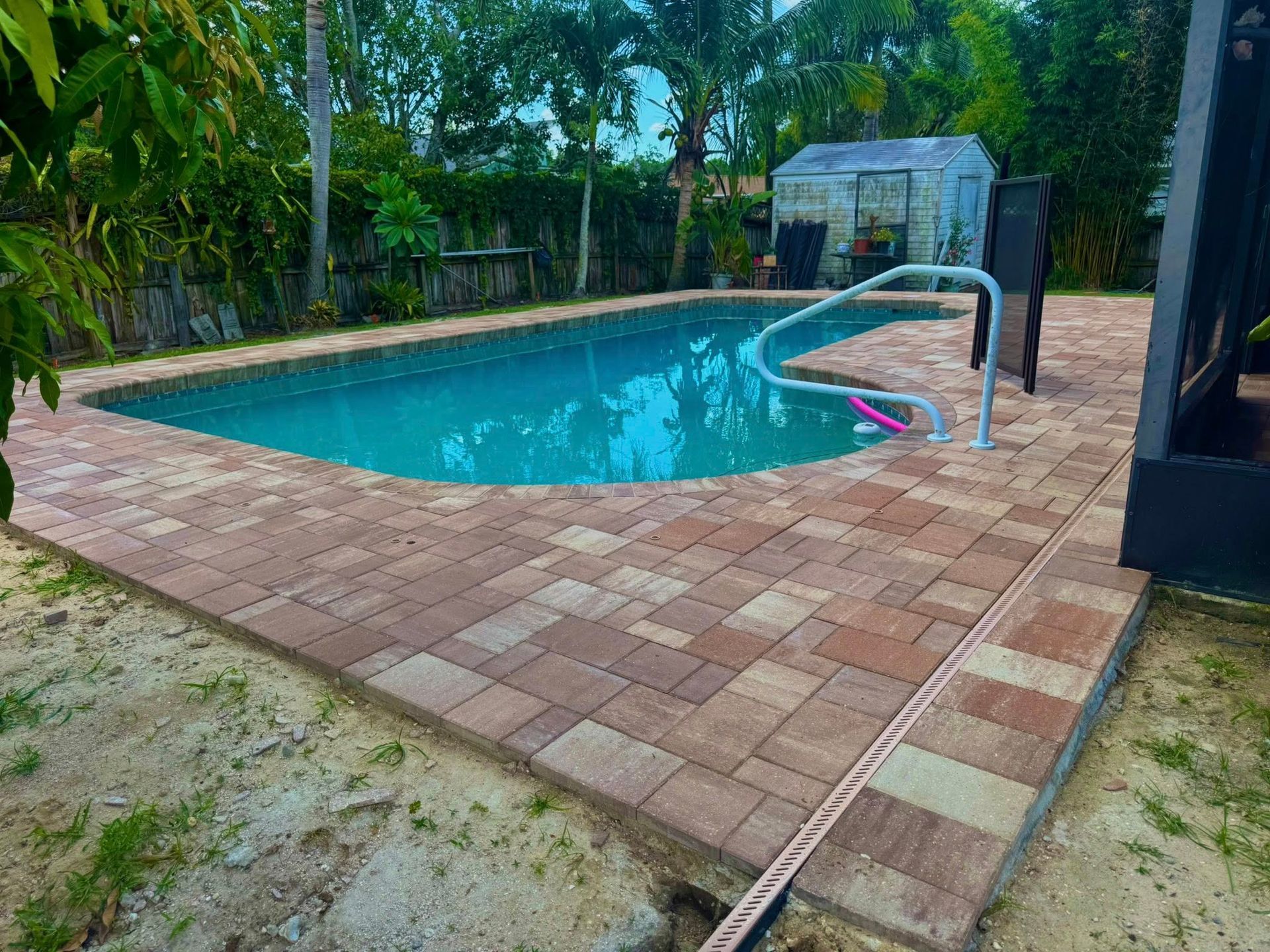 A rectangular pool with brick pavers, set in a yard. Green water, metal handrail, and small greenhouse are visible.