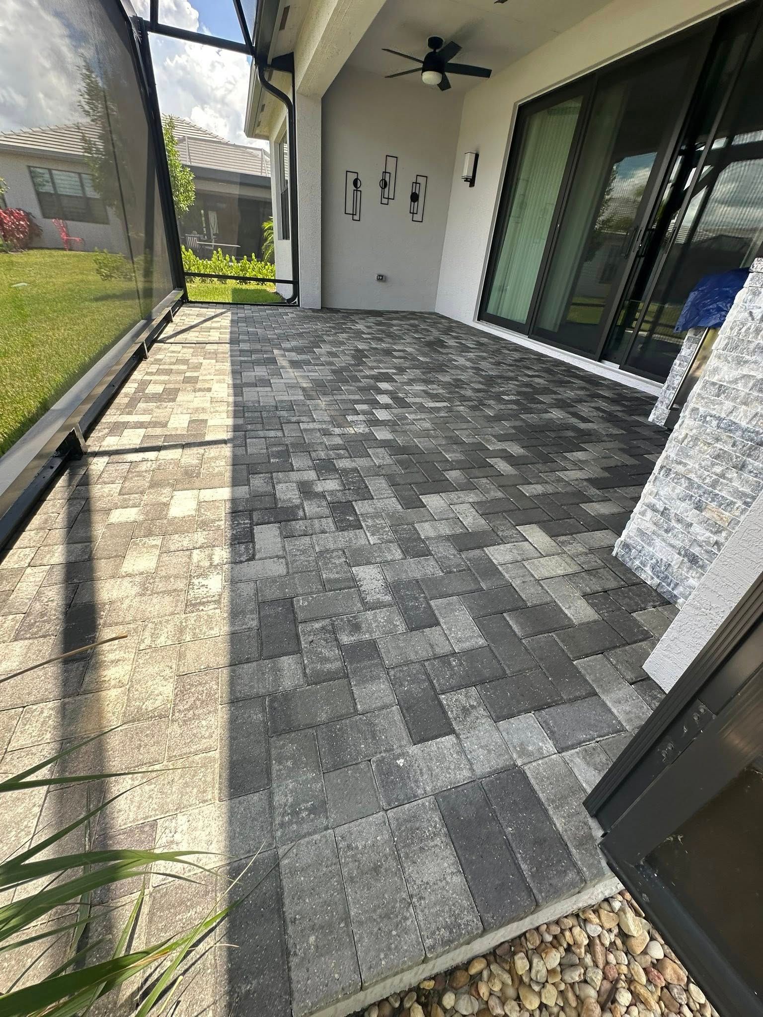 Patio with gray and black pavers, enclosed by a screen and connected to a house with sliding glass doors.