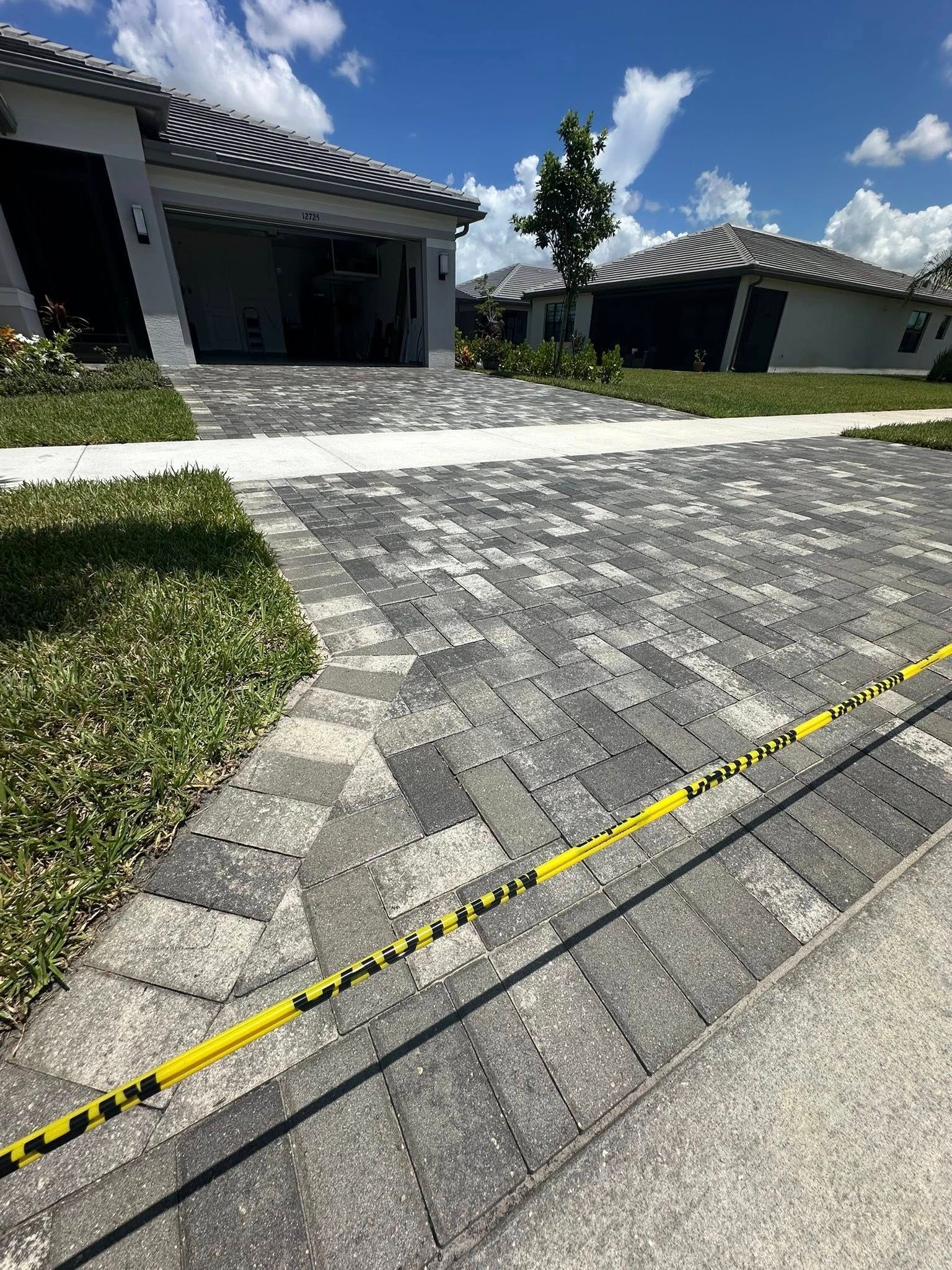 Driveway paved with gray and black bricks, caution tape, houses in the background, blue sky.