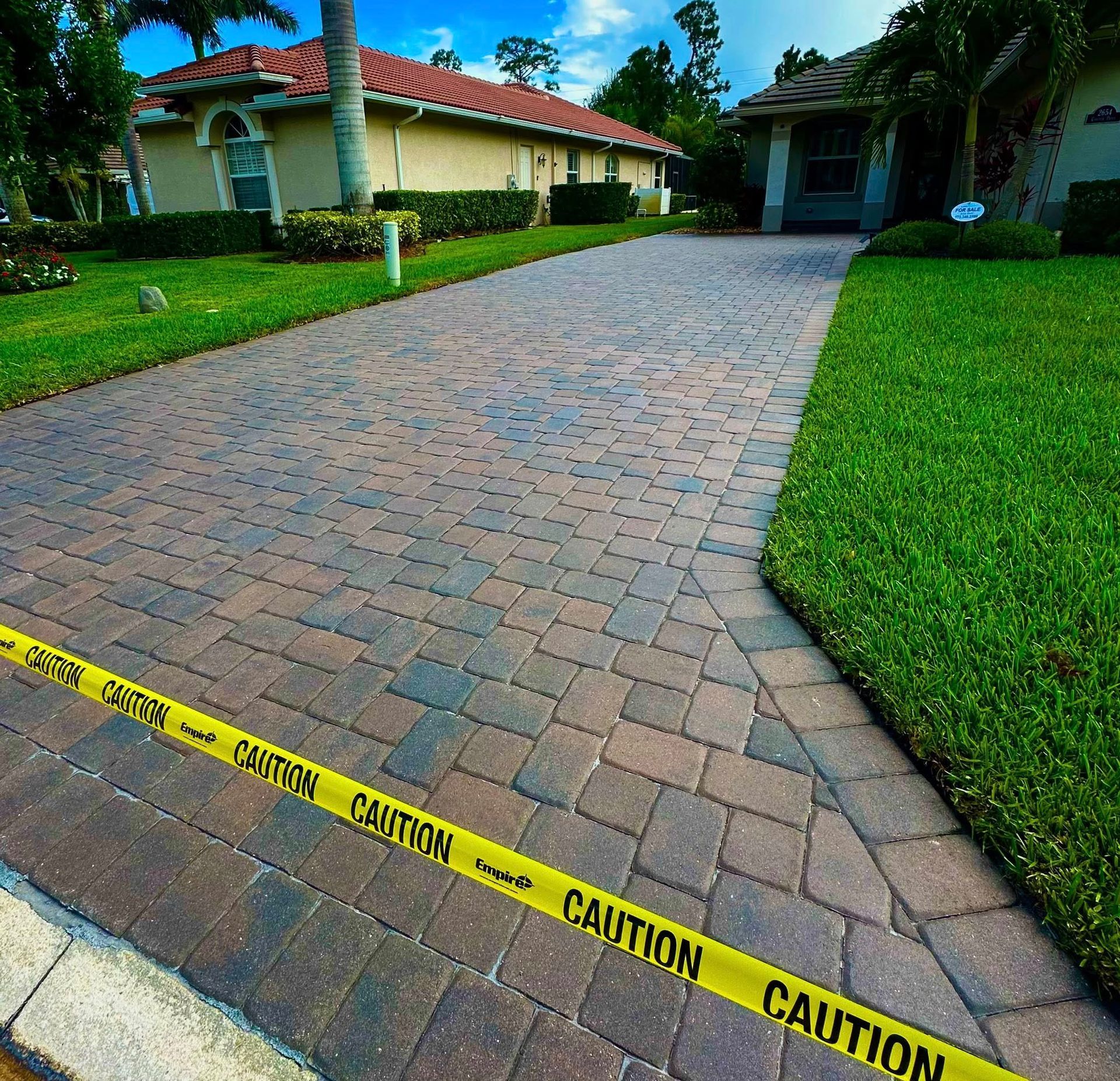 Driveway with cobblestone pavers, bordered by grass and caution tape. Houses in the background.