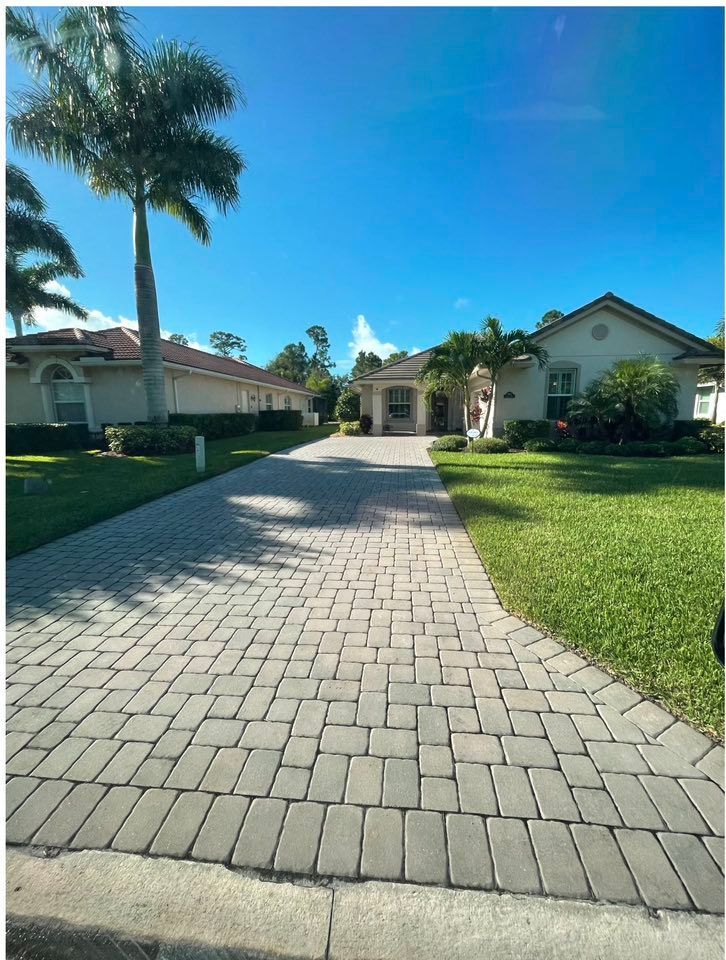 Brick driveway leads to suburban homes under a blue sky. Palm trees and green lawns.