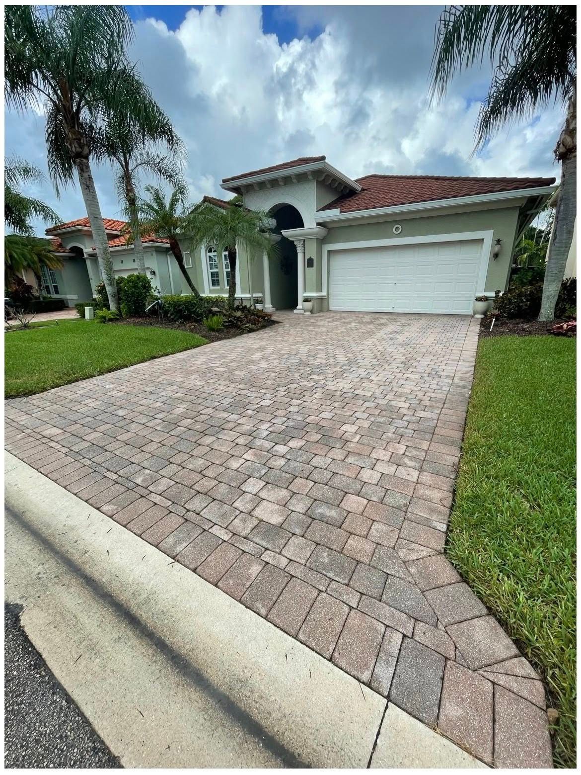 House with a brick driveway, green lawn, palm trees, and a white garage door against a cloudy blue sky.