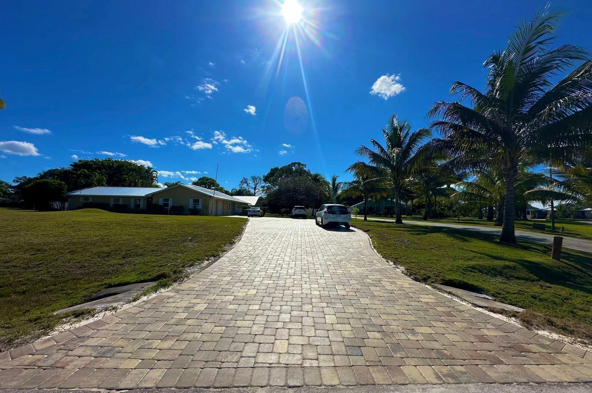 Brick driveway leading to houses on a sunny day with blue sky and palm trees.