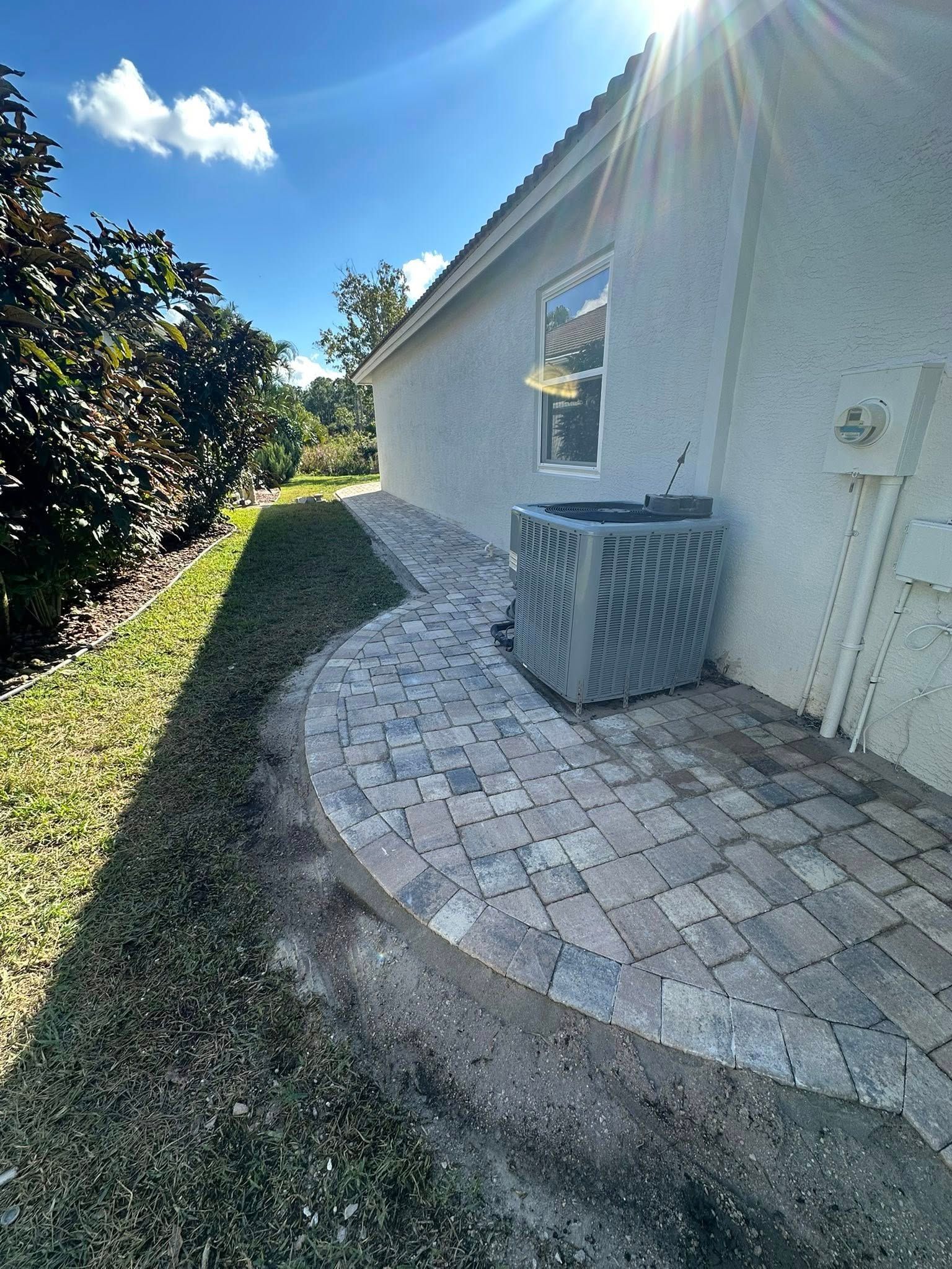 Exterior view of a house with a brick paved pathway and an air conditioning unit. Sunny day.