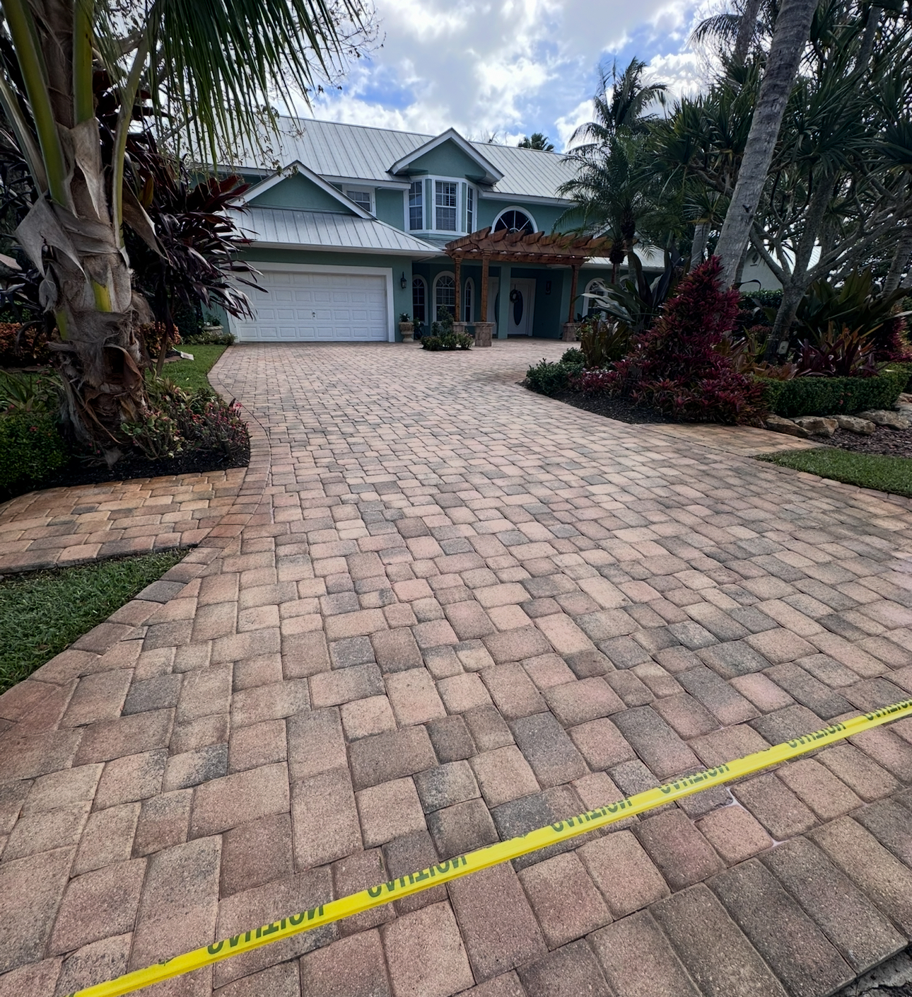 Driveway leading to a two-story blue house with a white garage door, palm trees, and brick pavers.