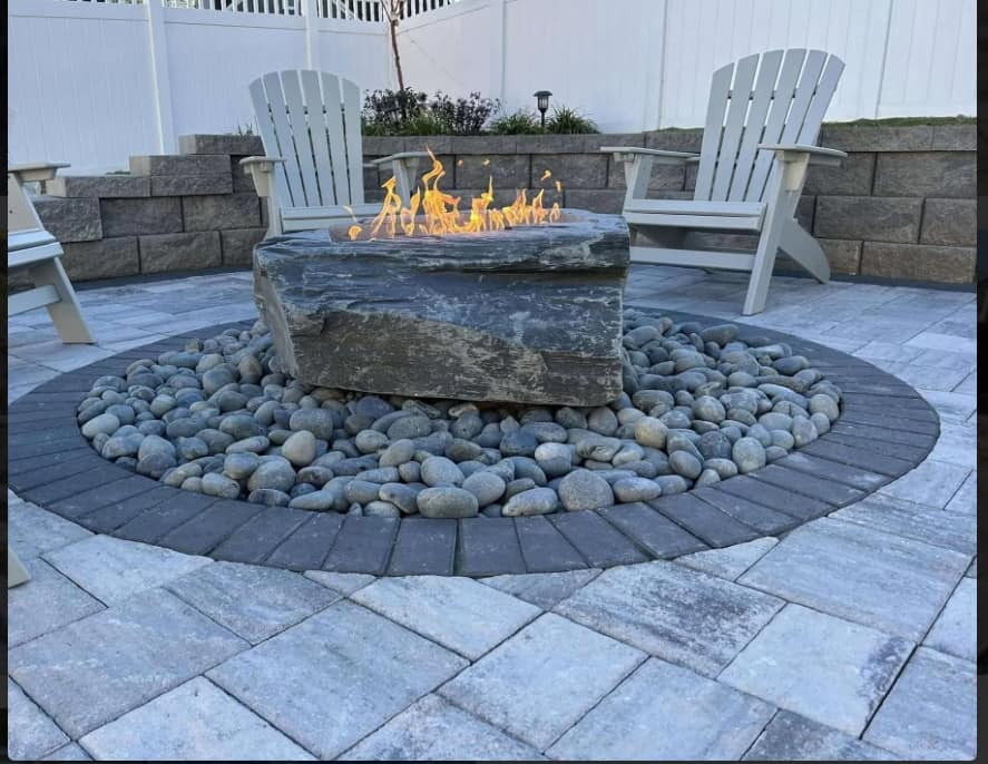 Outdoor fire pit with a large stone, surrounded by rocks and pavers, with Adirondack chairs.