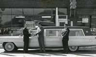 Three men shaking hands next to a long white hearse parked in front of a building.