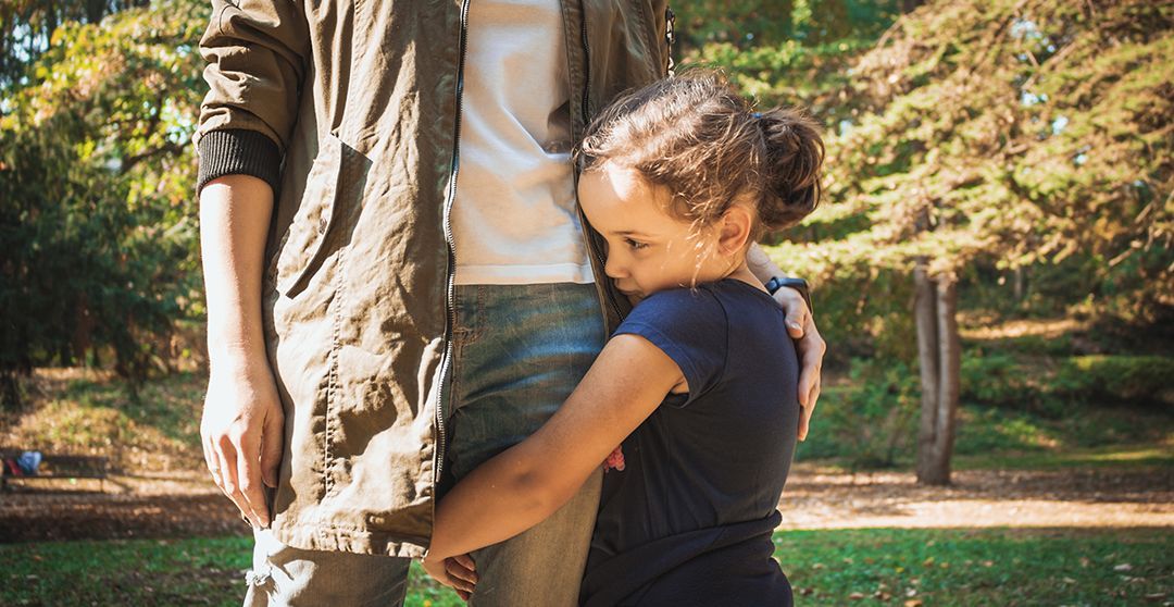 Girl hugging an adult's leg in a park; the girl has a worried expression.