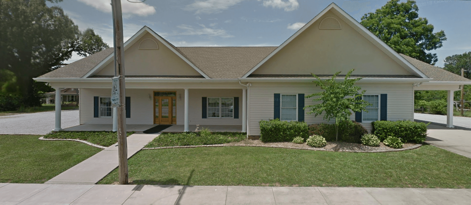 A one-story, light-colored building with a gray roof, green lawn, and bushes.