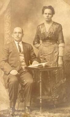 Older couple in formal attire, man seated with book, woman standing beside him. Studio portrait, sepia tones.