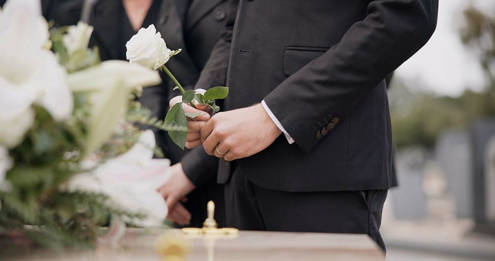 Man in black suit at a funeral, holding a white rose near a coffin and flowers.