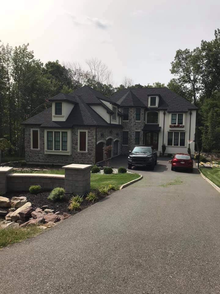 Large stone and white mansion with a long driveway. Two cars parked out front, with trees in the background.