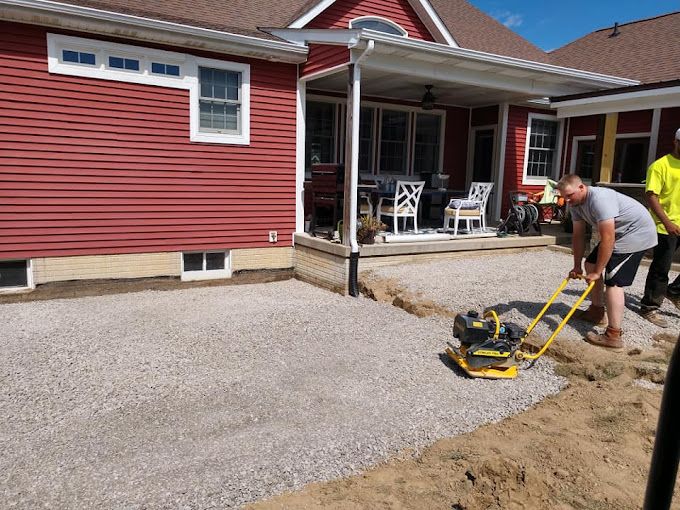 Two men are working on a driveway in front of a red house.