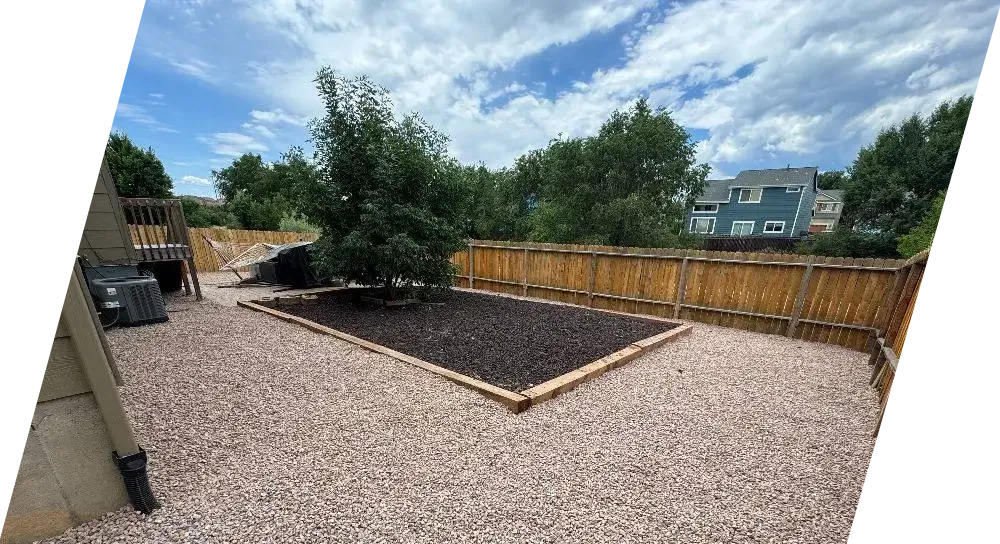 Backyard with gravel ground, a mulched bed with a tree, wooden fence, and a deck.