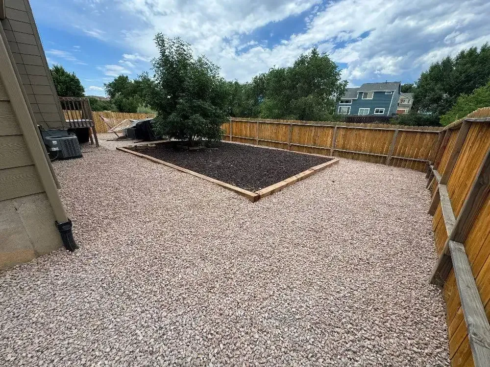 A backyard with a gravel ground, a fenced area, and a dark mulch garden bed with a small tree.