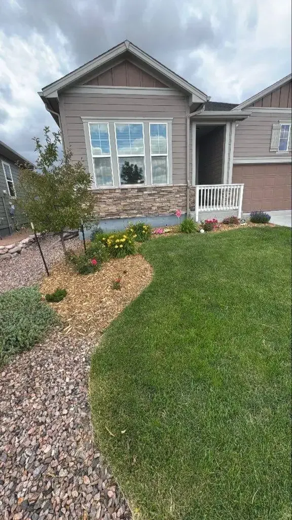 Tan house with front garden bed, flowers, and green lawn. Cloudy sky in background.