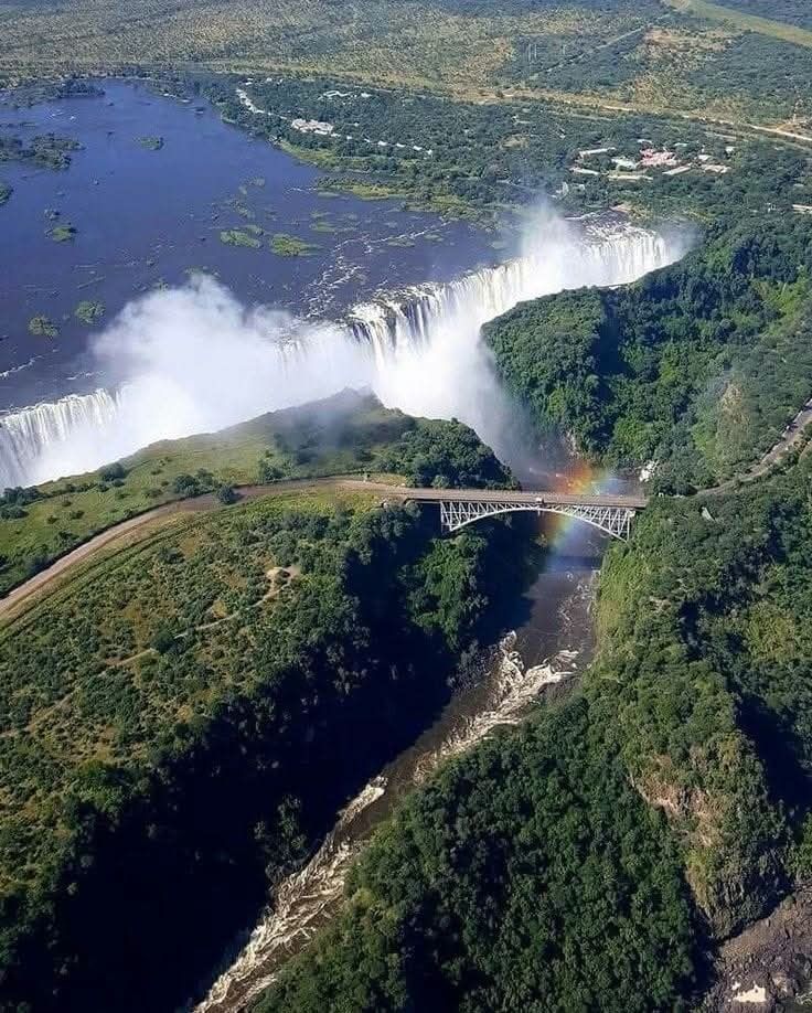 Victoria Falls, Zambia, seen from above. Rainbow arcs over the bridge spanning the Zambezi River. Water cascades over the falls.