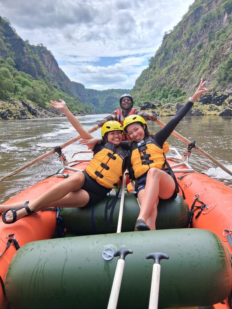 Three people on a raft in a river between cliffs, arms raised.