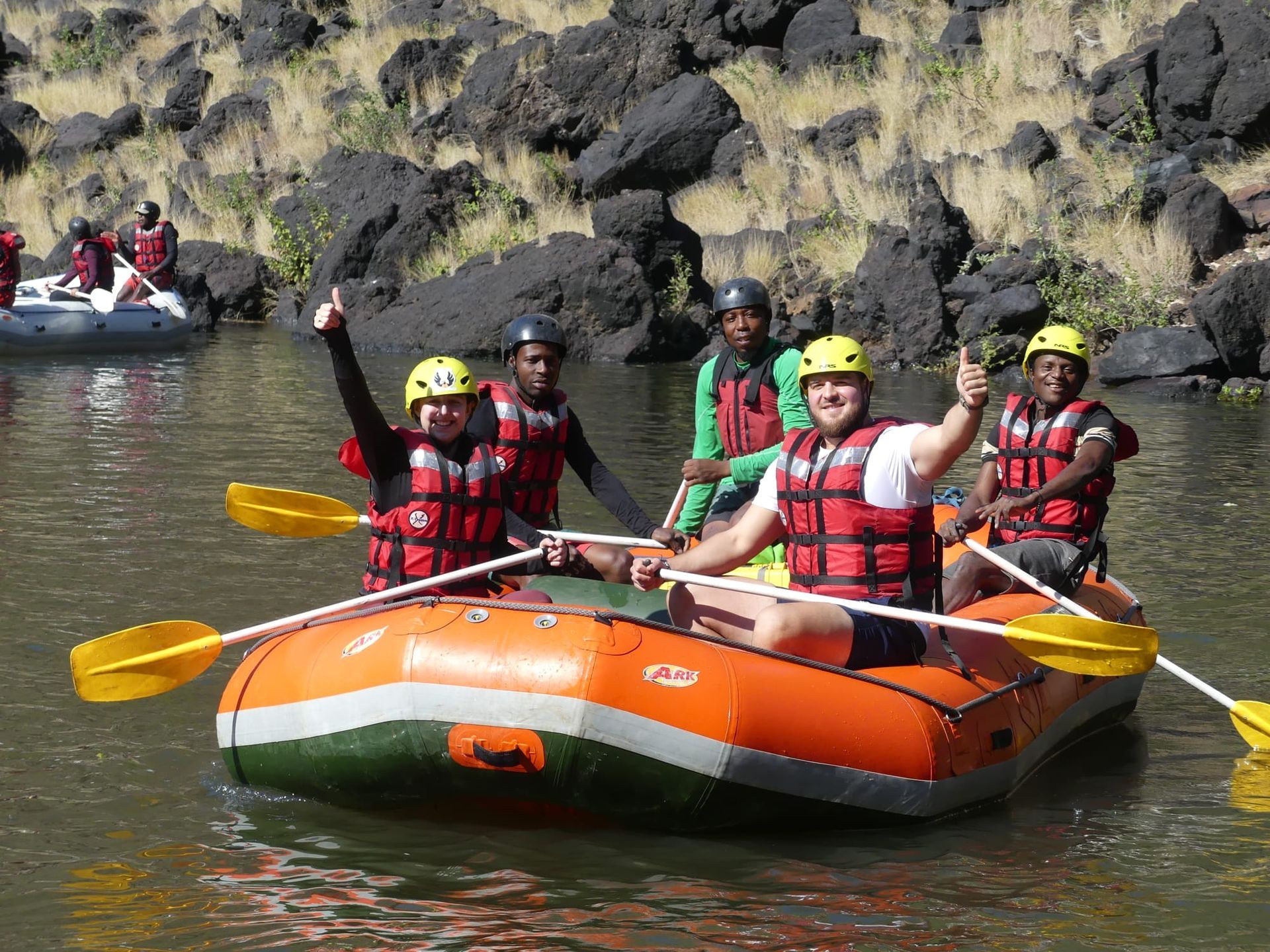 Rafting group in orange raft raises thumbs up. Green, brown water, black rocks in background.
