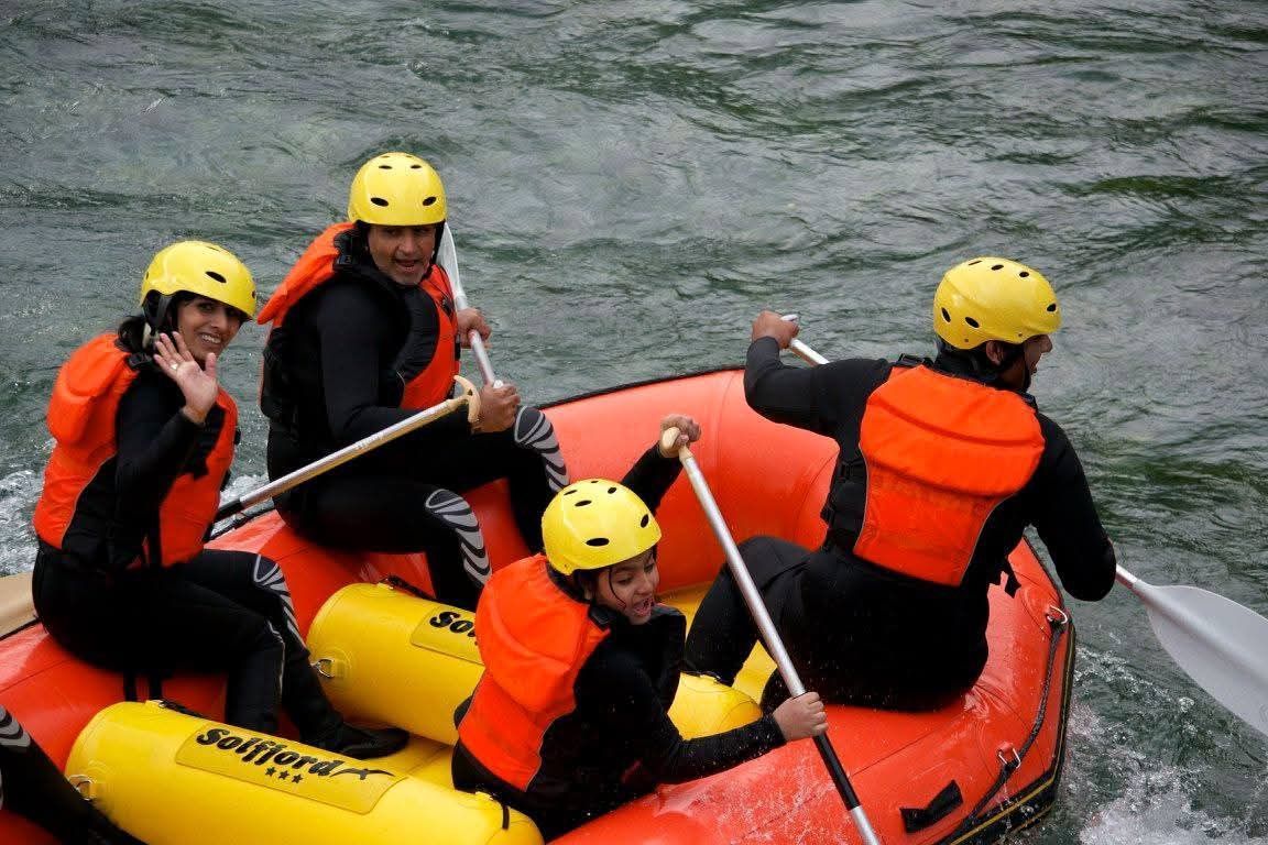 Four people in helmets and life vests raft on a river, paddling and smiling.