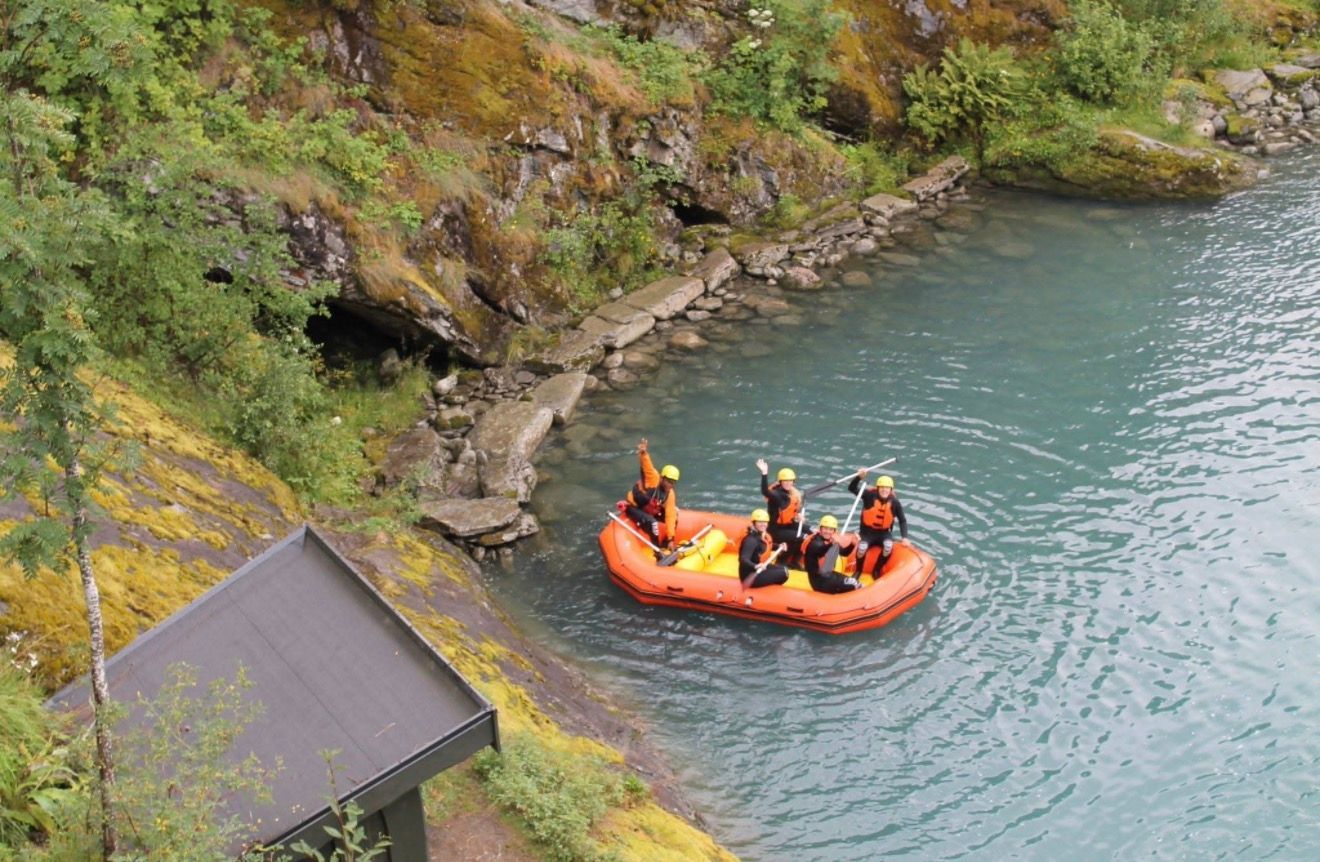 Orange raft with people in blue water near a rocky shore. Green moss, trees and small building in view