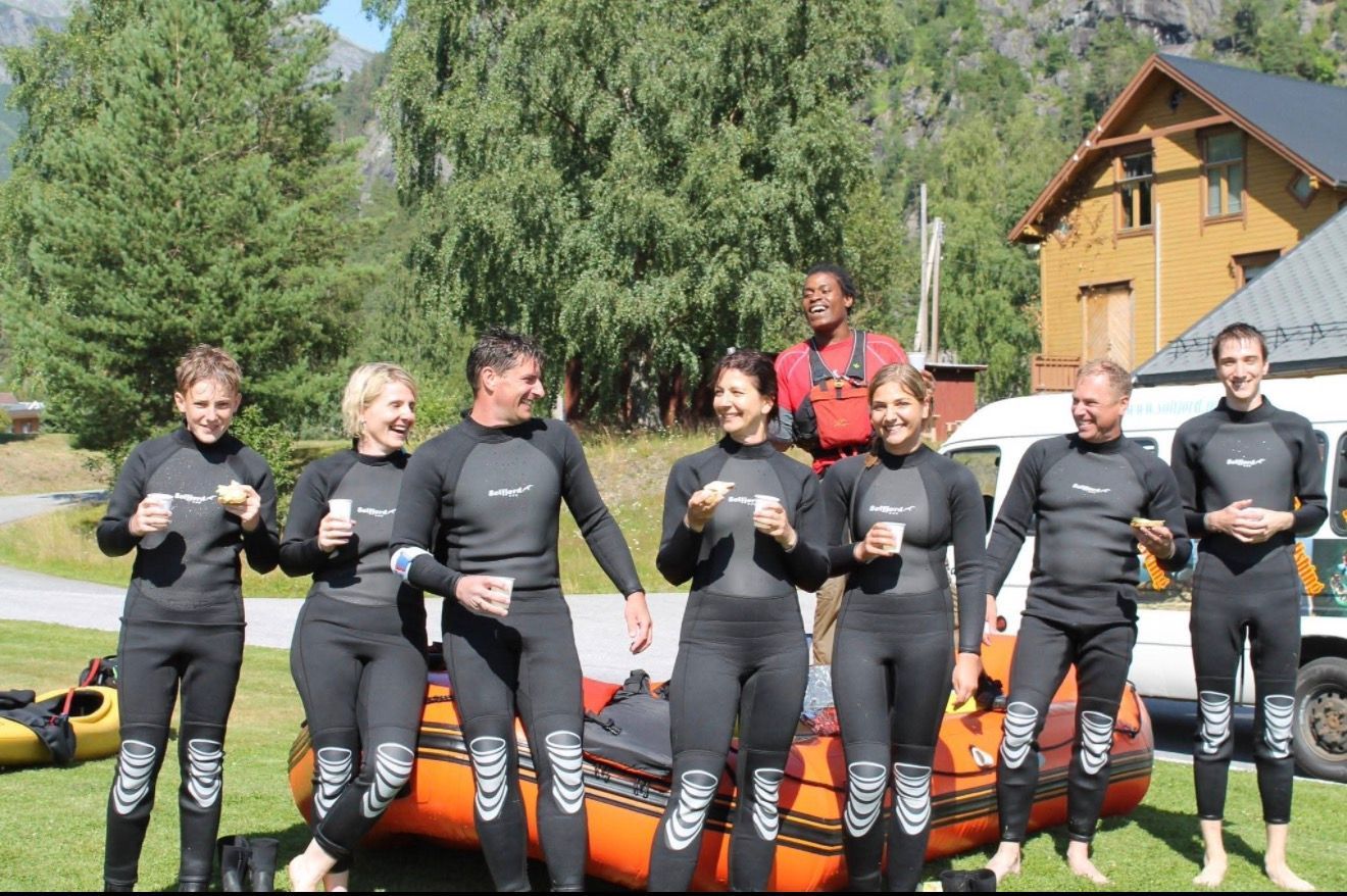 Group in wetsuits holding mugs, orange raft, standing by a building in a natural setting.