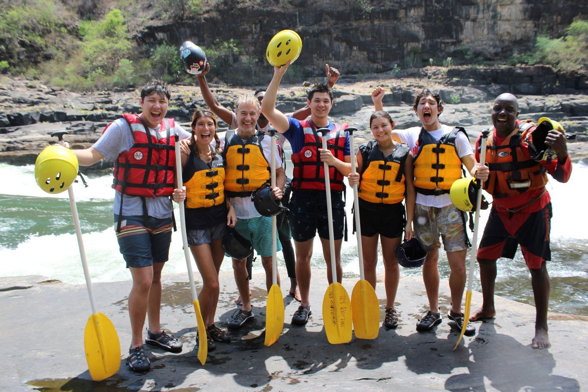 Group of people in life vests and helmets holding paddles, smiling and posing near a river.