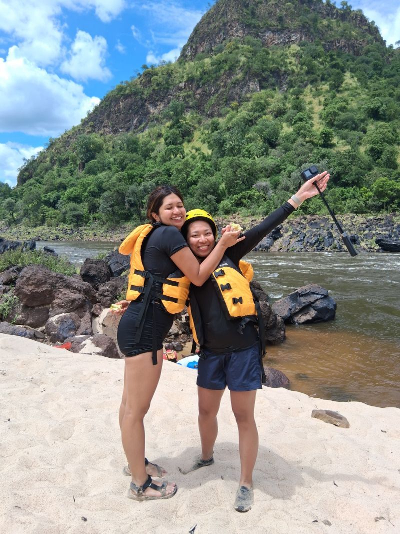 Two women are standing next to each other on a beach near a river.