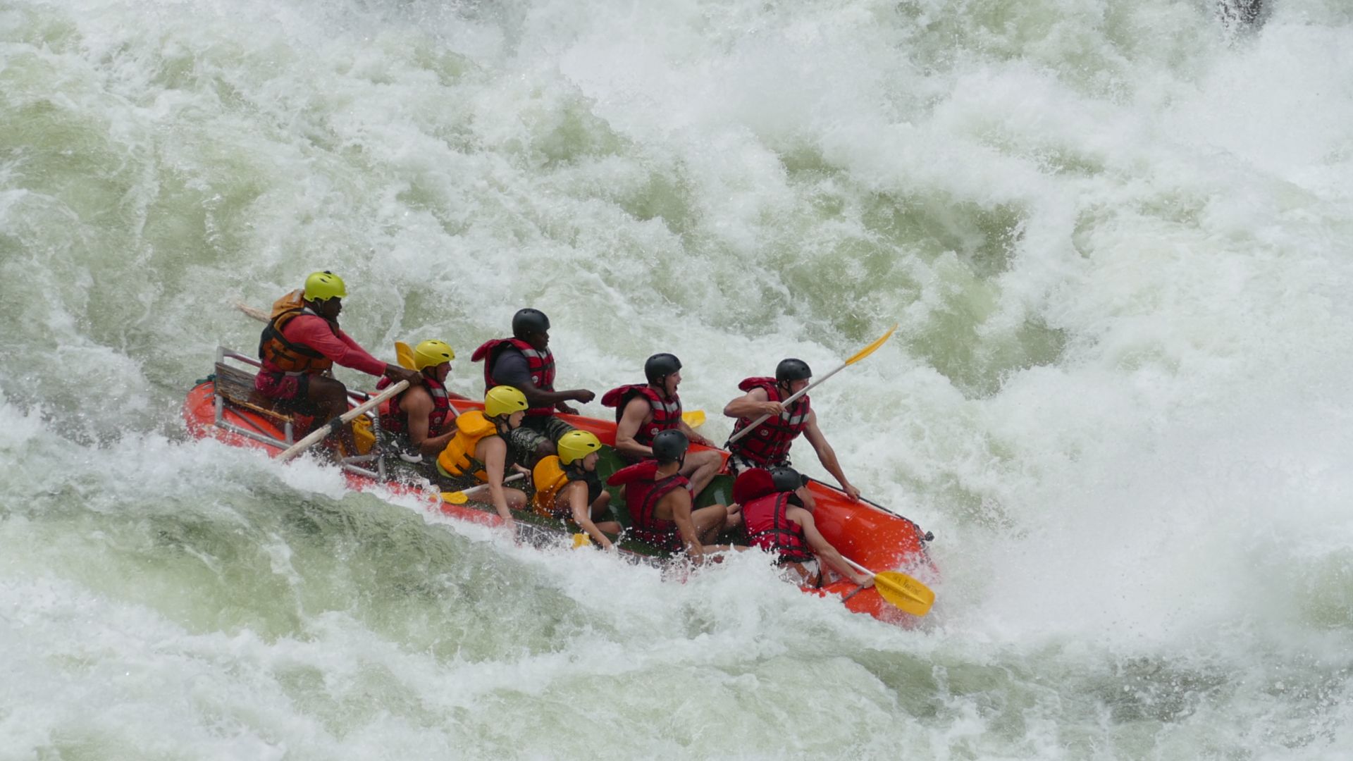 A group of people in life jackets and helmets paddle an orange raft through turbulent, white whitewater rapids.
