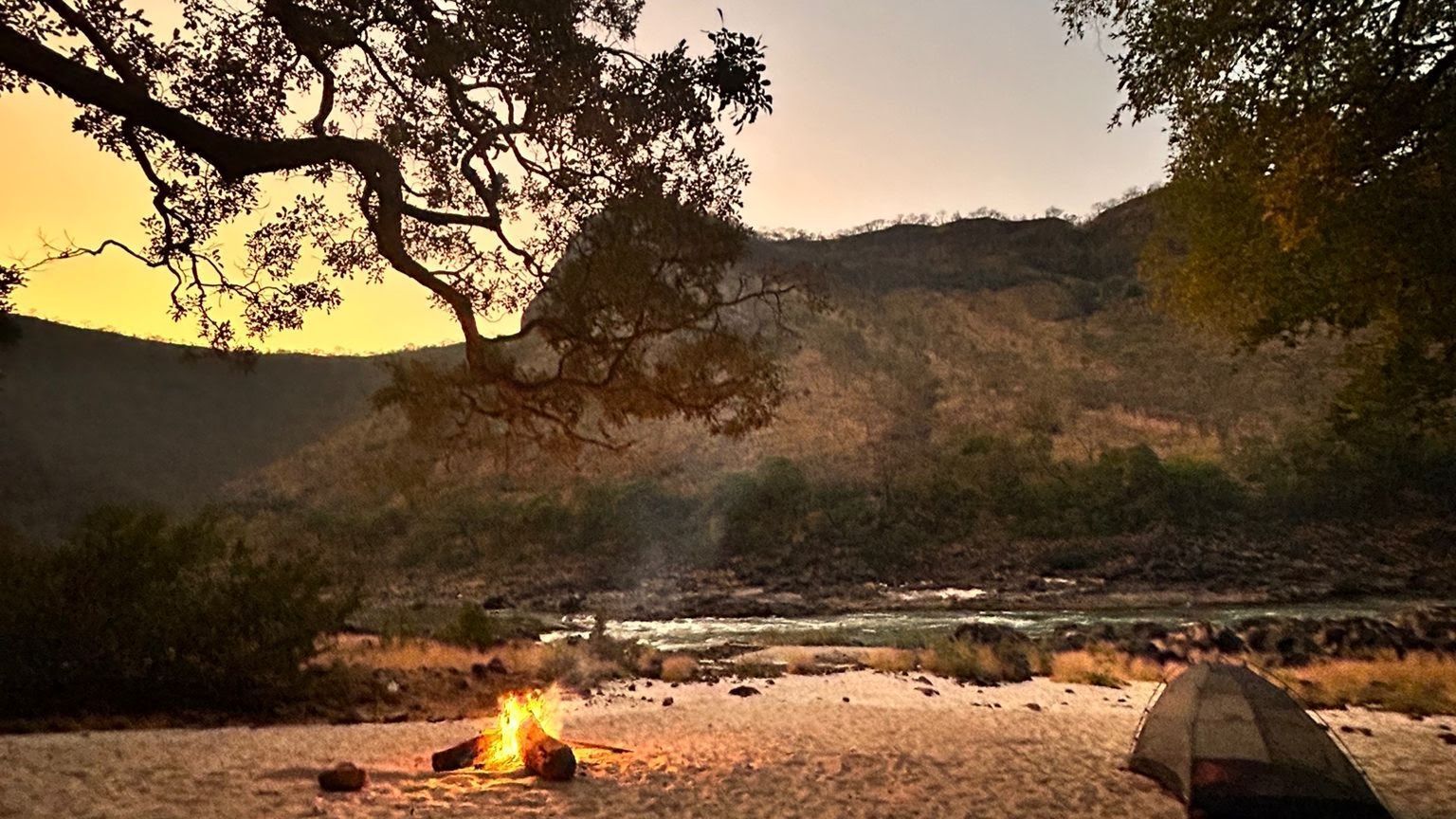 A tent is sitting next to a campfire on the shore of a river.